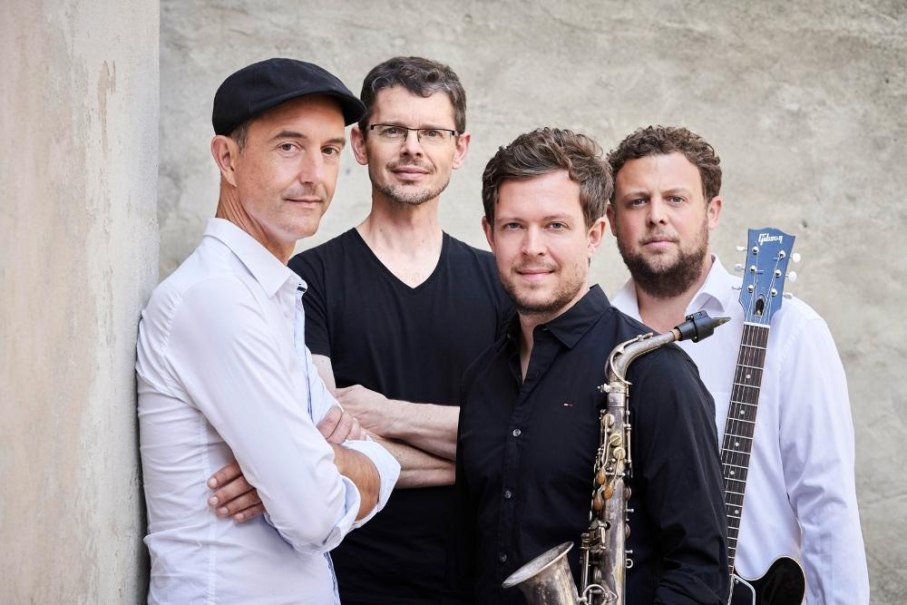 Vier M&auml;nner posieren vor einer Wand, einer mit Saxophon, ein anderer mit Gitarre. Sie tragen l&auml;ssige Kleidung und blicken in die Kamera., &copy; Foto Kaupo Kikkas