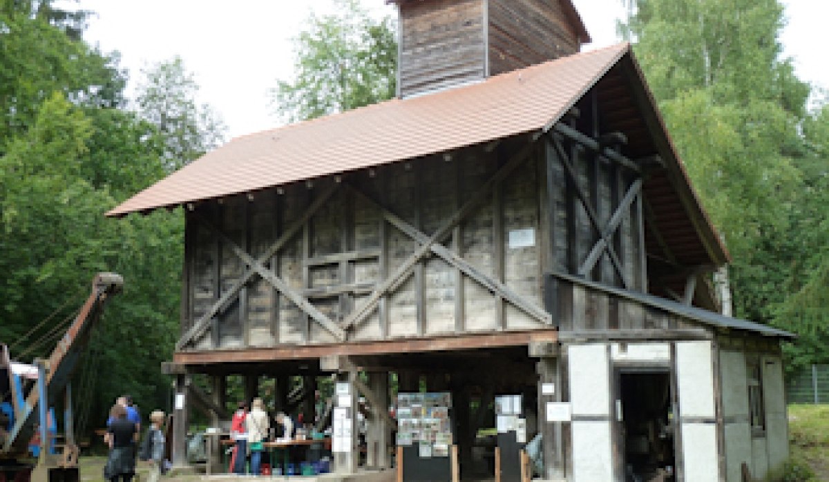 Historisches Sandwerk mit Holzkonstruktion und rotem Dach, umgeben von Bäumen. Menschen stehen davor und betrachten Ausstellungsstücke.