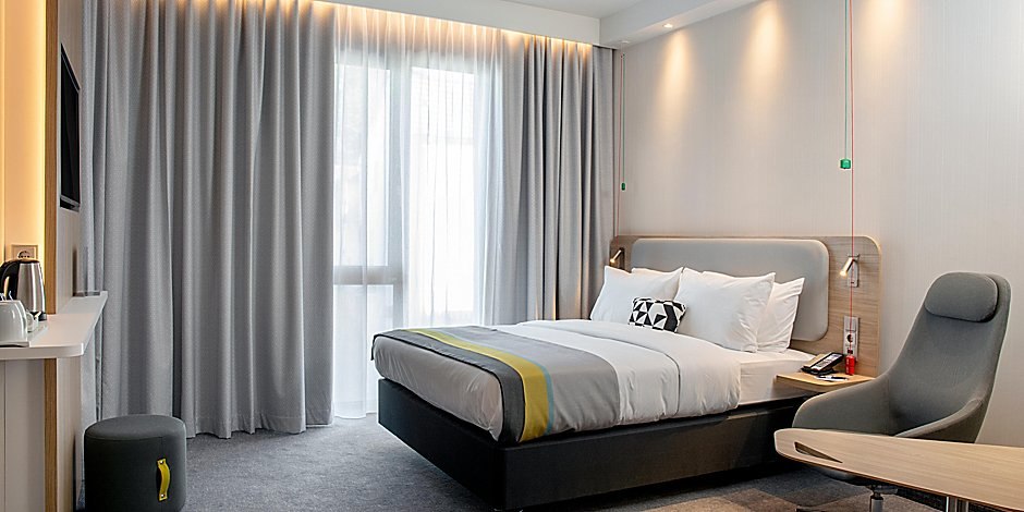 Modernes Hotelzimmer mit Doppelbett, grauen Vorhängen, Sessel und Schreibtisch. Dezente Beleuchtung und neutrale Farben dominieren den Raum., © Holiday Inn Express Waiblingen
