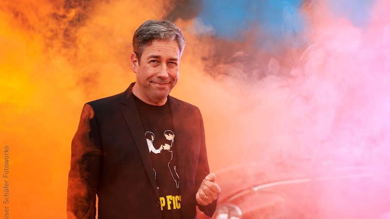 Ein Mann in einem schwarzen Anzug mit T-Shirt steht vor buntem Rauch in Orange und Blau. Im Hintergrund ist ein Auto zu erkennen., © Stadt Kornwestheim Kulturmanagement