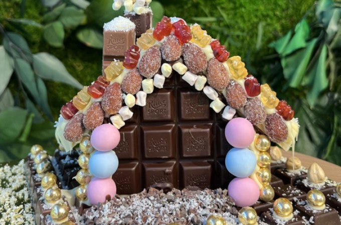 Ein mit Schokolade und bunten Süßigkeiten dekoriertes Lebkuchenhaus vor einem Hintergrund aus grünen Blättern., © Alfred Ritter GmbH &amp; Co. KG | Waldenbuch