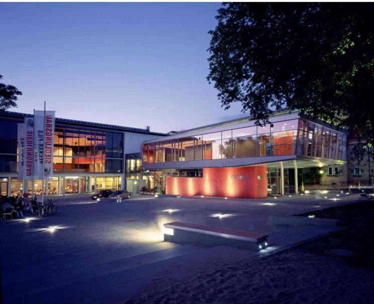 Die Stadthalle K3N in Nürtingen bei Dämmerung, modern beleuchtet mit Glasfassade und rotem Akzent. Fahrräder und Autos sind im Vordergrund sichtbar., © Peschke Die Stadthalle K3N in Nürtingen bei Dämmerung, modern beleuchtet mit Glasfassade und rotem Akzent. Fahrräder und Autos sind im Vordergrund sichtbar., © Peschke