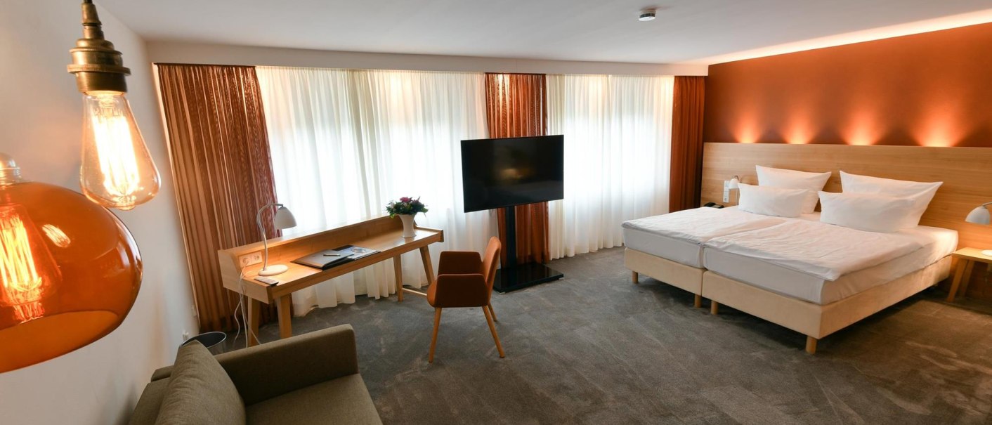 Modernes Hotelzimmer mit Doppelbett, Sofa, Schreibtisch und Flachbildfernseher. Warme Beleuchtung und Vorhänge schaffen eine gemütliche Atmosphäre., © Ringhotel Adler Asperg