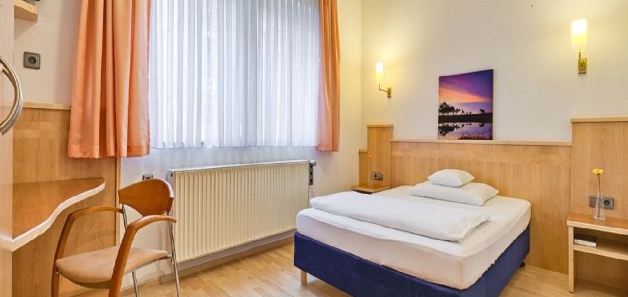Ein gemütliches Hotelzimmer mit Einzelbett, Holzmöbeln, Stuhl und Bild an der Wand. Große Fenster mit Vorhängen lassen Licht herein., © ARNOTEL GmbH Ein gemütliches Hotelzimmer mit Einzelbett, Holzmöbeln, Stuhl und Bild an der Wand. Große Fenster mit Vorhängen lassen Licht herein., © ARNOTEL GmbH