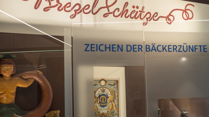 Ausstellung im Brezelmuseum mit dem Titel 'Brezelsch&auml;tze'. Zu sehen sind Exponate der B&auml;ckerz&uuml;nfte, darunter ein Schild und ein Gem&auml;lde., &copy; BrezelMuseum