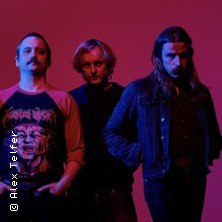 Drei M&auml;nner posieren vor einem roten Hintergrund. Einer tr&auml;gt ein T-Shirt mit einem Gesichtsmotiv. Die Stimmung ist d&uuml;ster und intensiv., &copy; links im Bild