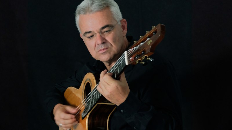 Ein Mann mit grauen Haaren spielt konzentriert auf einer Akustikgitarre vor einem dunklen Hintergrund., © Kultur- und Sportamt Stadt Bietigheim-Bissingen Ein Mann mit grauen Haaren spielt konzentriert auf einer Akustikgitarre vor einem dunklen Hintergrund., © Kultur- und Sportamt Stadt Bietigheim-Bissingen