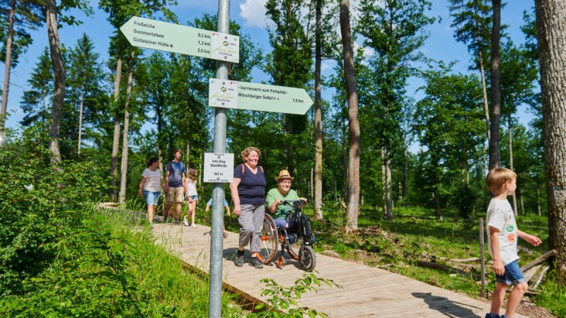 Ein Holzsteg im Wald mit mehreren Personen, darunter eine Person im Rollstuhl. Wegweiser zeigen Entfernungen zu verschiedenen Zielen an.