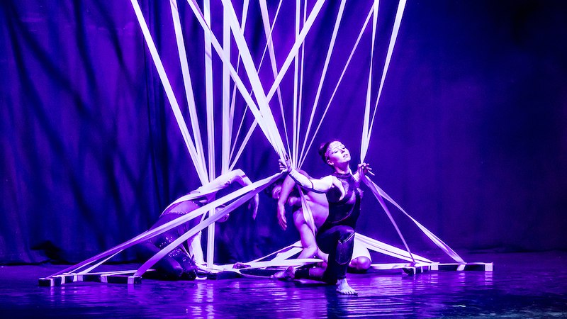 Zwei Tänzerinnen in violettem Licht, umgeben von weißen Bändern, auf einer Bühne. Eine kniet, die andere ist teils verdeckt. Dramatische Beleuchtung., © Stadthalle Leonberg