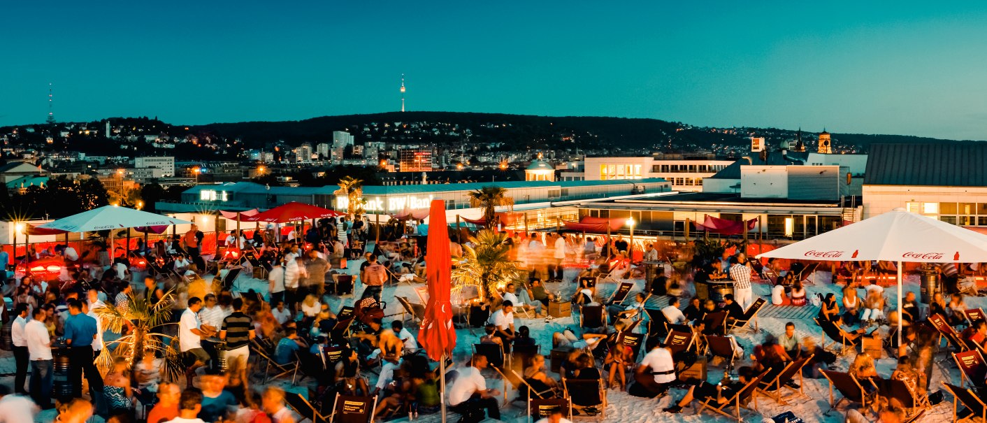 Große Menschenmenge auf einer Dachterrasse bei Abenddämmerung, umgeben von Sonnenschirmen und Liegestühlen, mit Blick auf eine beleuchtete Stadt., © Sky Beach