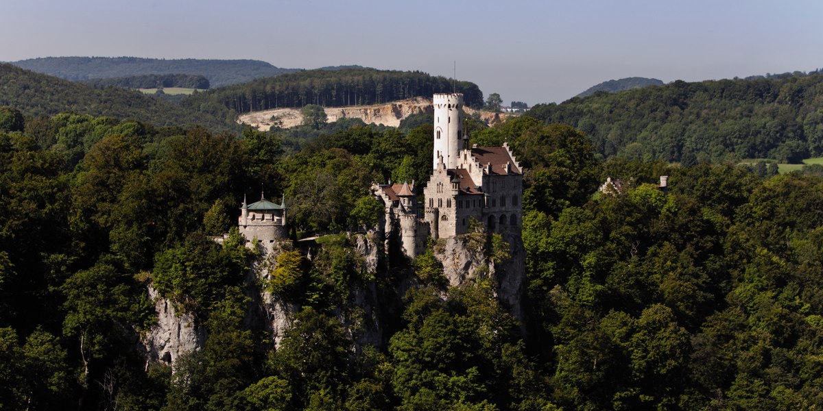 Schloss Lichtenstein thront majestätisch auf einem bewaldeten Hügel, umgeben von dichtem Wald und sanften Hügeln im Hintergrund., © Stuttgart-Marketing GmbH Schloss Lichtenstein thront majestätisch auf einem bewaldeten Hügel, umgeben von dichtem Wald und sanften Hügeln im Hintergrund., © Stuttgart-Marketing GmbH