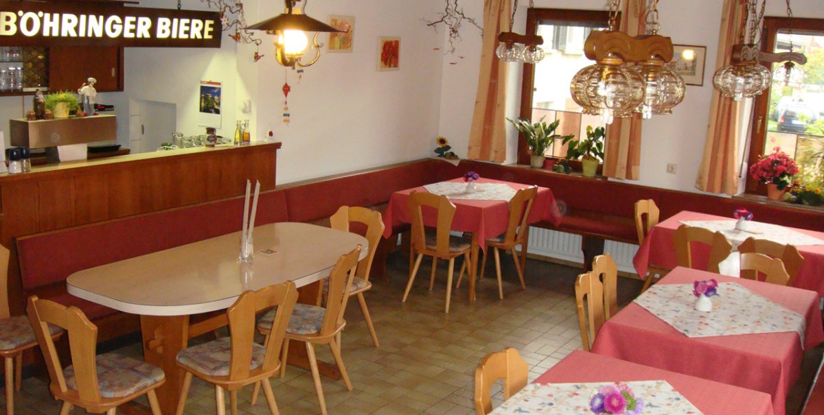 Ein gemütlicher Gastraum mit Holzmöbeln, roten Tischdecken und einer Bar mit "Böhringer Biere"-Schild. Pflanzen und Lampen dekorieren den Raum.