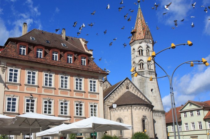 Historische Gebäude mit einem Kirchturm im Hintergrund, Tauben fliegen am blauen Himmel. Menschen sitzen unter Sonnenschirmen., © Touristik und Marketing GmbH