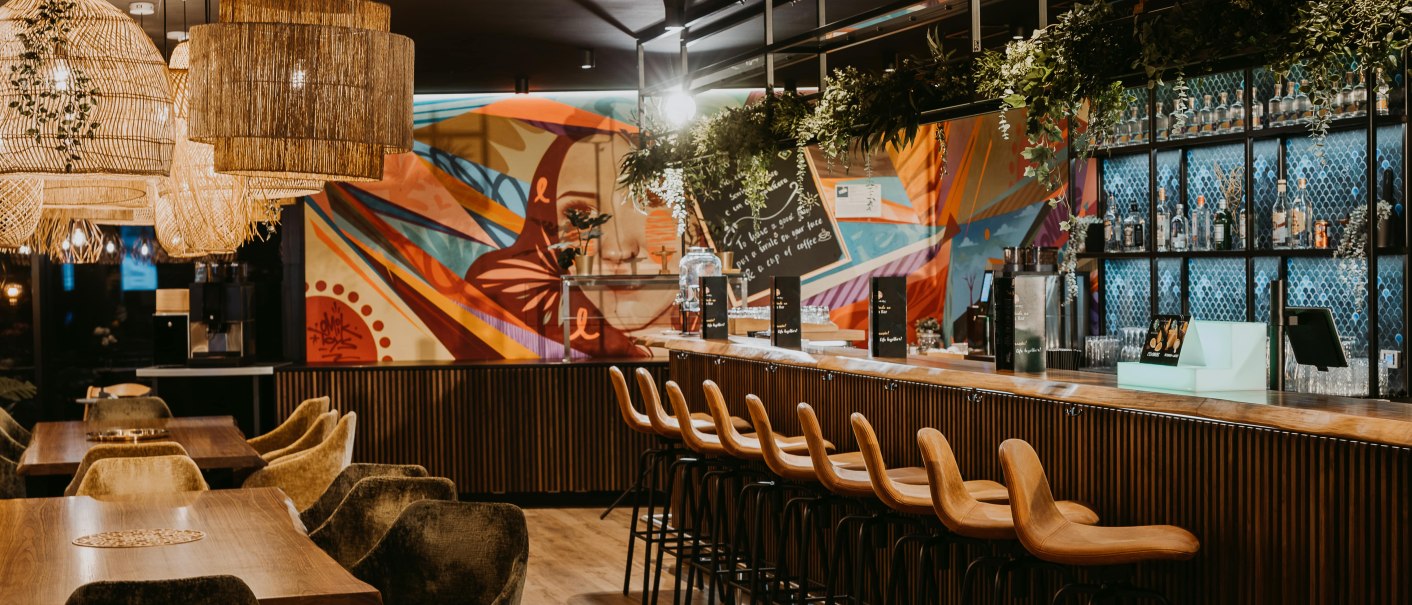 Moderne Bar mit Holztresen, hohen Stühlen und buntem Wandgemälde. Hängelampen und Pflanzen dekorieren den Raum., © Rioca Stuttgart Posto 4 GmbH