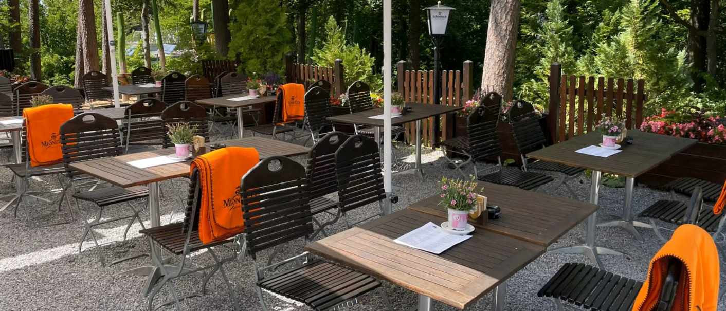 Ein ruhiger Biergarten mit Holztischen und -stühlen, orange Decken und Pflanzen auf den Tischen, umgeben von Bäumen., © Schönbuch Waldbiergarten am Wasserturm Ein ruhiger Biergarten mit Holztischen und -stühlen, orange Decken und Pflanzen auf den Tischen, umgeben von Bäumen., © Schönbuch Waldbiergarten am Wasserturm