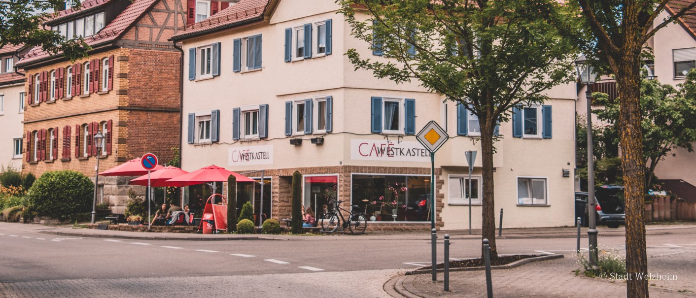 Ein Fachwerkhaus und ein Café mit roten Sonnenschirmen an einer Straßenecke in einer deutschen Stadt, umgeben von Bäumen., © Stadt Welzheim