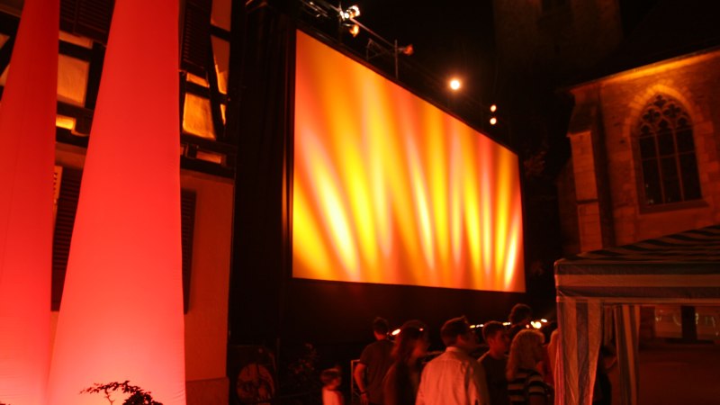 Freiluftkino bei Nacht mit gro&szlig;er, beleuchteter Leinwand. Menschen stehen davor, umgeben von warmem Licht und Geb&auml;uden im Hintergrund., &copy; Torsten Wenzler