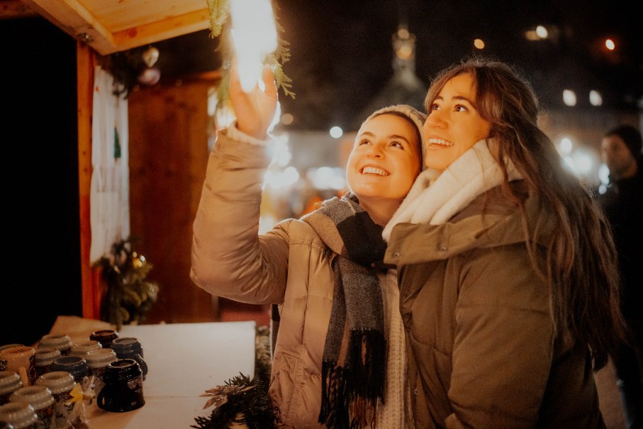 Zwei Frauen stehen auf einem Weihnachtsmarkt und schauen lächelnd nach oben. Sie tragen warme Kleidung und sind von Lichtern umgeben., © Nik Ebert