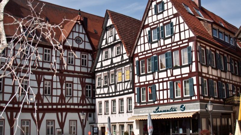 Fachwerkhäuser mit roten Dächern und blauen Fensterläden. Vor dem Wirtshaus Laurentia sitzen Menschen im Freien bei sonnigem Wetter. Fachwerkhäuser mit roten Dächern und blauen Fensterläden. Vor dem Wirtshaus Laurentia sitzen Menschen im Freien bei sonnigem Wetter.