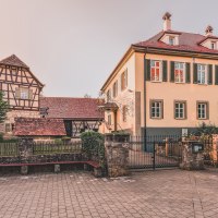 Ein historisches Fachwerkhaus neben einem modernen Gebäude mit grünen Fensterläden in Welzheim. Davor ein gepflasterter Platz und ein schmiedeeisernes Tor., © Stadt Welzheim Ein historisches Fachwerkhaus neben einem modernen Gebäude mit grünen Fensterläden in Welzheim. Davor ein gepflasterter Platz und ein schmiedeeisernes Tor., © Stadt Welzheim