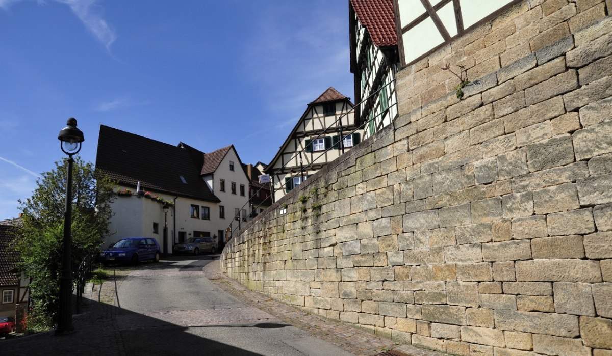 Gepflasterte Straße mit Fachwerkhäusern und einer Steinmauer, blauer Himmel, Laterne und Autos im Hintergrund., © Natur.Nah. Schönbuch & Heckengäu Gepflasterte Straße mit Fachwerkhäusern und einer Steinmauer, blauer Himmel, Laterne und Autos im Hintergrund., © Natur.Nah. Schönbuch & Heckengäu