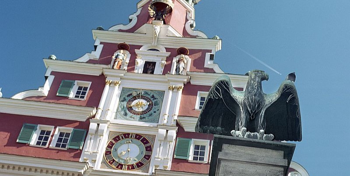 Das Bild zeigt das Alte Rathaus mit seiner markanten Uhr und einer Adlerstatue im Vordergrund, unter einem klaren blauen Himmel.