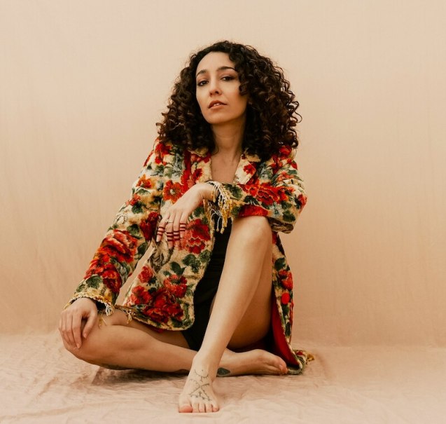 Eine Frau mit lockigem Haar sitzt in einem geblümten Mantel auf einem beigen Hintergrund. Sie wirkt entspannt und selbstbewusst., © Kulturhaus Schwanen