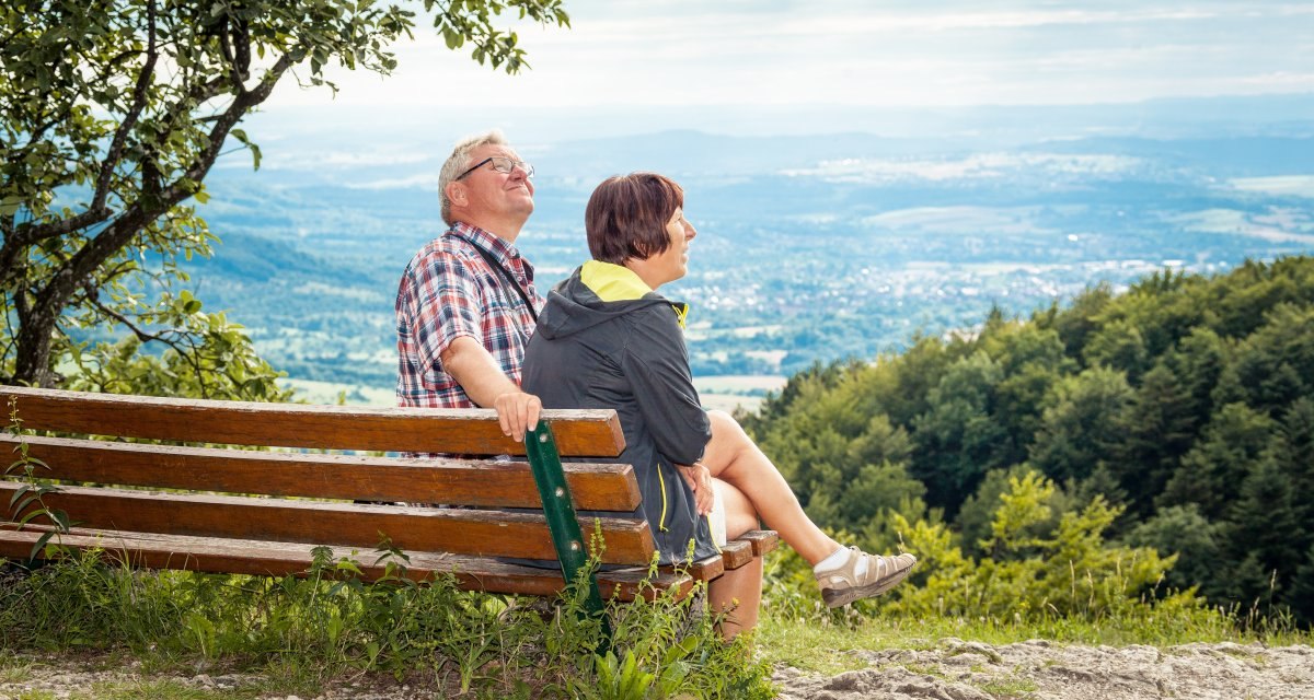 Ein älteres Paar sitzt auf einer Bank und genießt die Aussicht auf eine weite, grüne Landschaft unter einem bewölkten Himmel., © hochgehberge Ein älteres Paar sitzt auf einer Bank und genießt die Aussicht auf eine weite, grüne Landschaft unter einem bewölkten Himmel., © hochgehberge