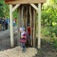 Kinder spielen in einer Holzkonstruktion aus Holzstämmen im Wald. Im Hintergrund stehen Erwachsene und beobachten die Szene., © Amt für Forsten Landkreis Böblingen