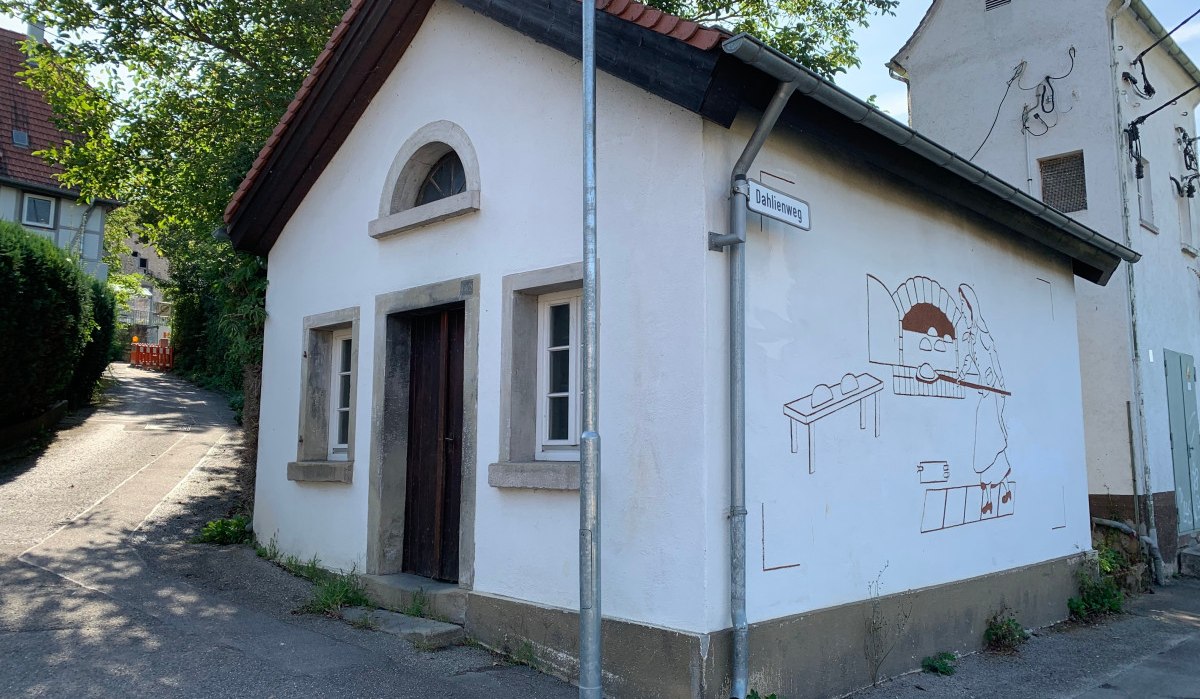Kleines weißes Gebäude mit rotem Dach, Wandgemälde eines Bäckers, Straßenschild 'Dahlienweg', Bäume im Hintergrund., © Land der 1000 Hügel - Kraichgau-Stromberg Kleines weißes Gebäude mit rotem Dach, Wandgemälde eines Bäckers, Straßenschild 'Dahlienweg', Bäume im Hintergrund., © Land der 1000 Hügel - Kraichgau-Stromberg