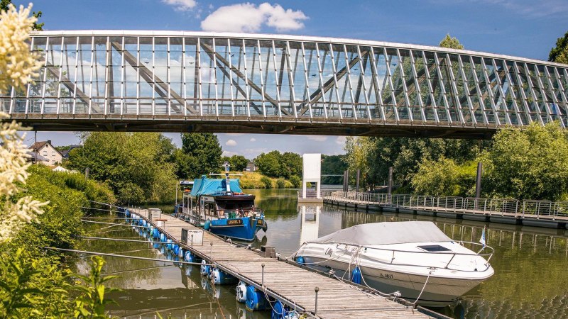 Moderne Glasbrücke über einem Fluss mit Booten und blühenden Pflanzen. Der Himmel ist klar und blau, was eine friedliche Sommeratmosphäre schafft., © Stuttgart-Marketing GmbH, Sarah Schmid Moderne Glasbrücke über einem Fluss mit Booten und blühenden Pflanzen. Der Himmel ist klar und blau, was eine friedliche Sommeratmosphäre schafft., © Stuttgart-Marketing GmbH, Sarah Schmid