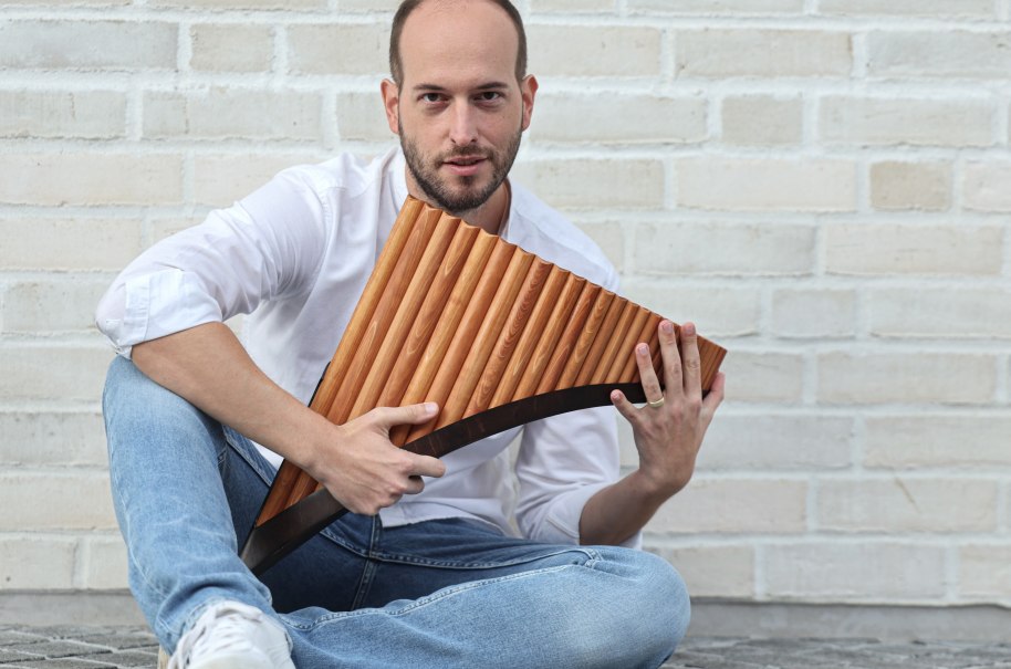 Ein Mann in wei&szlig;em Hemd und Jeans sitzt vor einer wei&szlig;en Backsteinwand und h&auml;lt eine Panfl&ouml;te in den H&auml;nden., &copy; Kevin Sch&auml;fer