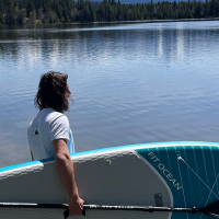 Eine Person steht am Ufer eines Sees mit einem SUP-Board und Paddel, umgeben von Wald und ruhigem Wasser., © seezeit GmbH