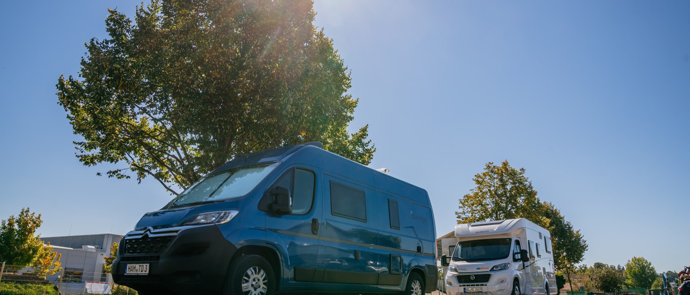Zwei Wohnmobile stehen auf einem sonnigen Stellplatz. Ein blauer Van und ein weißes Wohnmobil sind zu sehen, umgeben von Bäumen und blauem Himmel., © Stuttgart-Marketing GmbH, Thomas Niedermüller