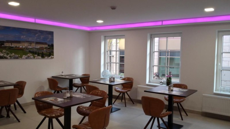 Moderner Loungebereich mit lila Beleuchtung, Tischen und Stühlen. An der Wand hängt ein großes Bild, Fenster lassen Tageslicht herein., © TOMAS Moderner Loungebereich mit lila Beleuchtung, Tischen und Stühlen. An der Wand hängt ein großes Bild, Fenster lassen Tageslicht herein., © TOMAS