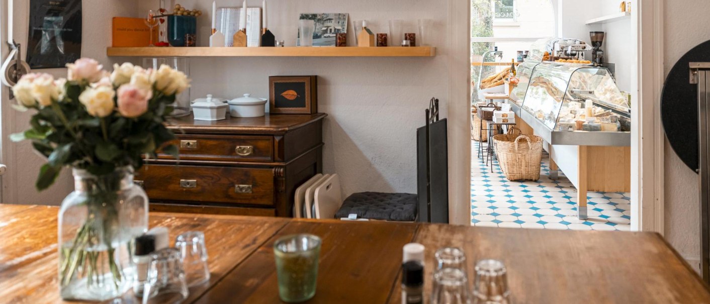 Ein gemütlicher Feinkostladen mit Holztisch, Blumenstrauß und einer Käsetheke im Hintergrund auf blau-weißem Fliesenboden., © SMG, Sarah Schmid Ein gemütlicher Feinkostladen mit Holztisch, Blumenstrauß und einer Käsetheke im Hintergrund auf blau-weißem Fliesenboden., © SMG, Sarah Schmid