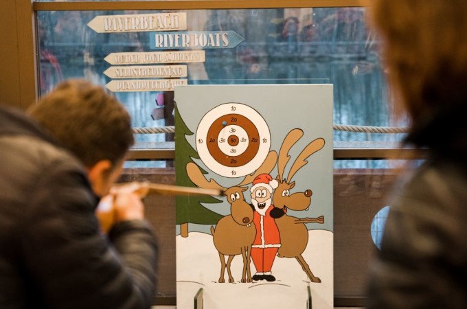 Zwei Personen zielen mit Blasrohren auf eine Zielscheibe mit einem Weihnachtsmann und Rentieren. Im Hintergrund sind Schilder und ein Fenster zu sehen., © Cool-Tours StattReisen