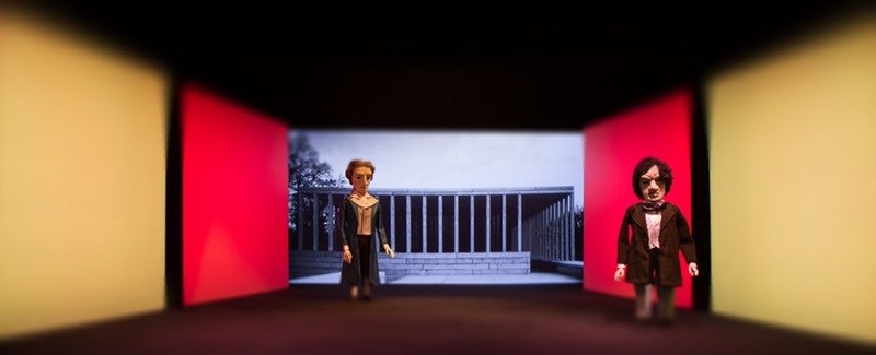 Zwei Puppen auf einer Bühne mit roten und gelben Wänden. Im Hintergrund ein schwarz-weißes Bild eines Gebäudes mit Säulen., © LIMA-Theater Zwei Puppen auf einer Bühne mit roten und gelben Wänden. Im Hintergrund ein schwarz-weißes Bild eines Gebäudes mit Säulen., © LIMA-Theater