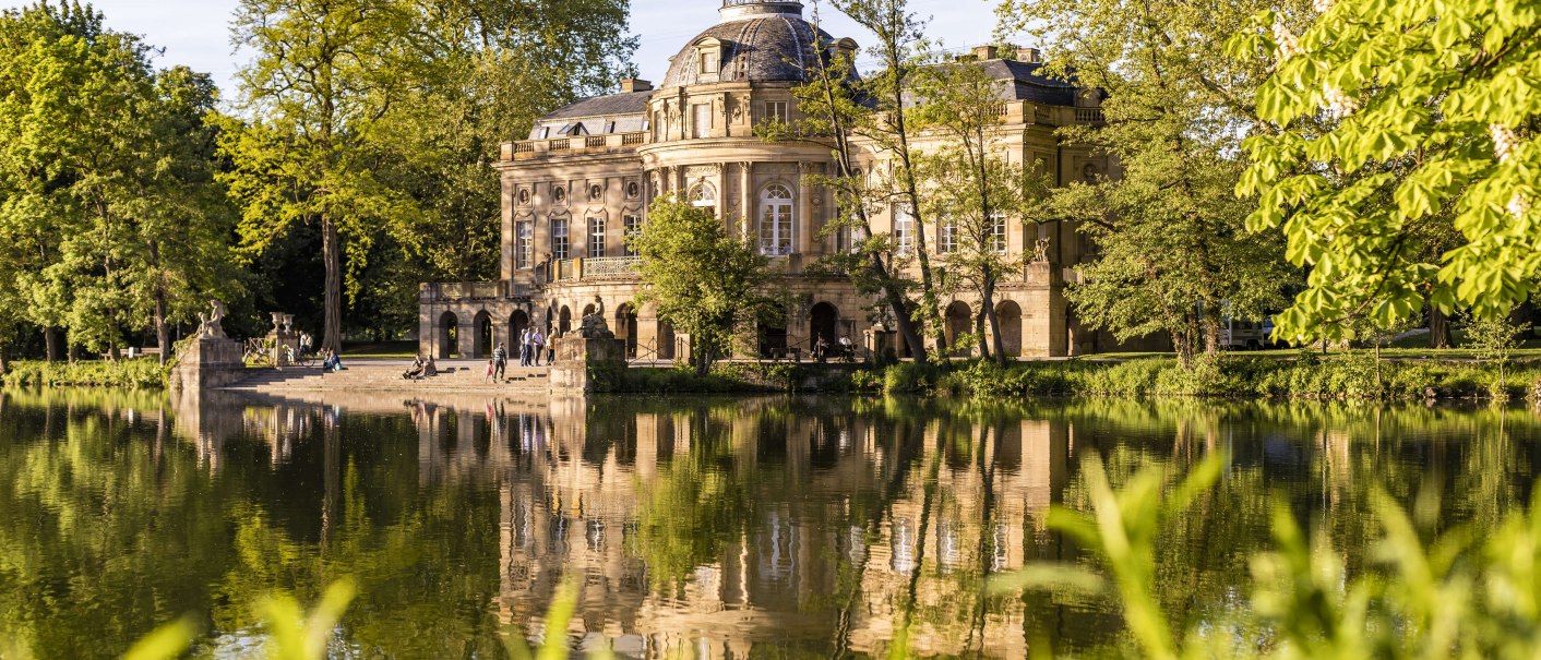 Schloss Monrepos spiegelt sich im ruhigen Wasser eines Sees, umgeben von üppigem Grün und Bäumen. Menschen spazieren am Ufer entlang., © SMG, Werner Dieterich Schloss Monrepos spiegelt sich im ruhigen Wasser eines Sees, umgeben von üppigem Grün und Bäumen. Menschen spazieren am Ufer entlang., © SMG, Werner Dieterich