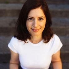 Eine Frau mit braunen Haaren und weißem T-Shirt sitzt lächelnd auf einer Treppe., © links im Bild Eine Frau mit braunen Haaren und weißem T-Shirt sitzt lächelnd auf einer Treppe., © links im Bild