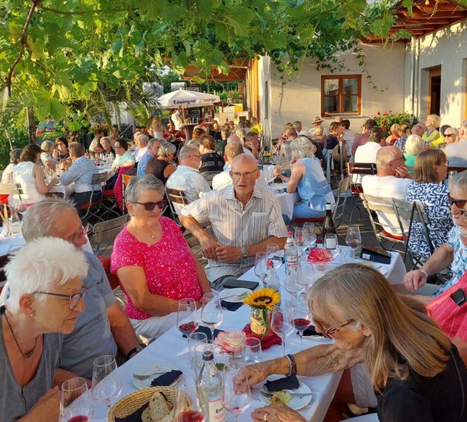Geselliges Beisammensein im Freien unter Weinreben. Menschen sitzen an Tischen, genie&szlig;en Getr&auml;nke und unterhalten sich in entspannter Atmosph&auml;re., &copy; Weingut Bayer