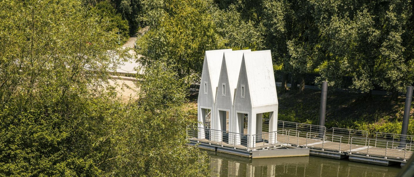 Ein modernes Badehaus mit spitzen Dächern steht auf einem Fluss, umgeben von üppigem Grün. Es ist über einen Steg erreichbar., © SMG, Sarah Schmid Ein modernes Badehaus mit spitzen Dächern steht auf einem Fluss, umgeben von üppigem Grün. Es ist über einen Steg erreichbar., © SMG, Sarah Schmid
