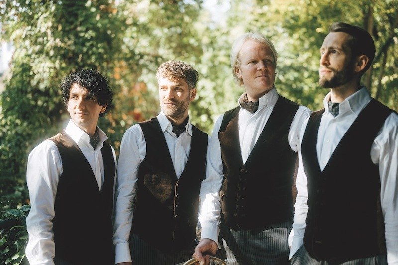 Vier M&auml;nner in eleganter Kleidung stehen im Freien und blicken in verschiedene Richtungen. Sie tragen Westen und Hemden, umgeben von gr&uuml;ner Natur., &copy; Kulturstiftung Hohenlohe