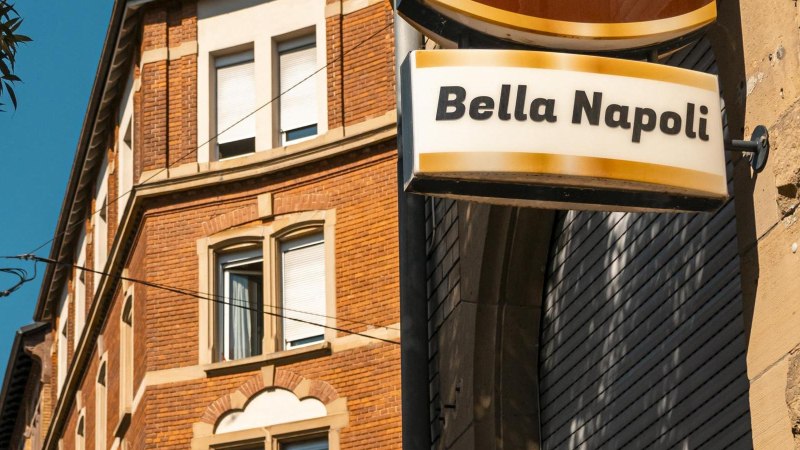 Ein Schild mit der Aufschrift 'Bella Napoli' h&auml;ngt an einem Backsteingeb&auml;ude. Der Himmel ist klar und blau, ein Baum ist im Vordergrund sichtbar., &copy; SMG, Sarah Schmid
