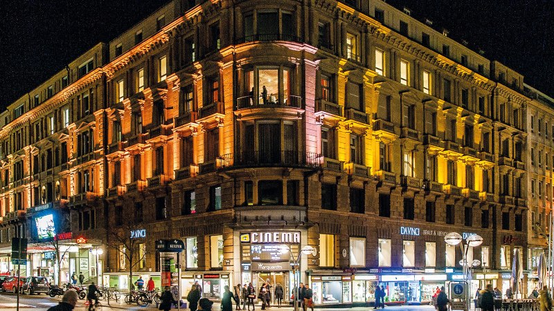 Ein großes, beleuchtetes Gebäude bei Nacht mit Geschäften im Erdgeschoss und Menschen auf der Straße., © Schauspielbühnen in Stuttgart Ein großes, beleuchtetes Gebäude bei Nacht mit Geschäften im Erdgeschoss und Menschen auf der Straße., © Schauspielbühnen in Stuttgart