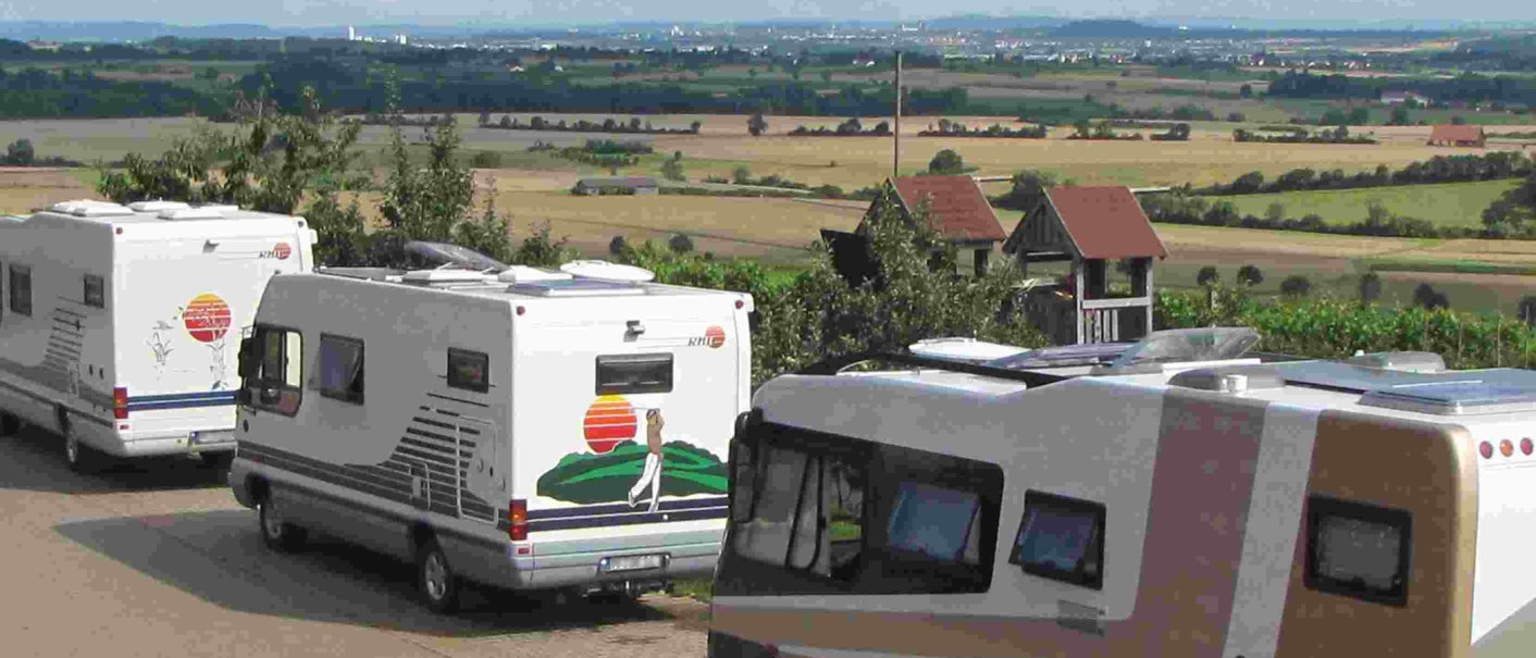 Wohnmobile parken auf einem Stellplatz mit Blick auf eine weite Landschaft und Felder. Im Hintergrund sind Hügel und eine Stadt zu sehen., © PANORAMAWEINGUT Baumgärtner Wohnmobile parken auf einem Stellplatz mit Blick auf eine weite Landschaft und Felder. Im Hintergrund sind Hügel und eine Stadt zu sehen., © PANORAMAWEINGUT Baumgärtner