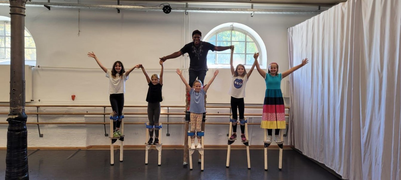 Kinder und ein Erwachsener stehen auf Stelzen in einem Raum. Sie halten sich an den Händen und lächeln. Im Hintergrund sind Vorhänge und Fenster zu sehen., © Kunstschule Labyrinth Kinder und ein Erwachsener stehen auf Stelzen in einem Raum. Sie halten sich an den Händen und lächeln. Im Hintergrund sind Vorhänge und Fenster zu sehen., © Kunstschule Labyrinth