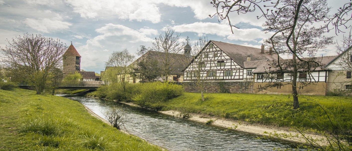 Fachwerkhäuser und ein Turm am Flussufer, umgeben von Bäumen und einer Brücke. Der Himmel ist bewölkt, die Landschaft wirkt idyllisch., © SMG, Sarah Schmid