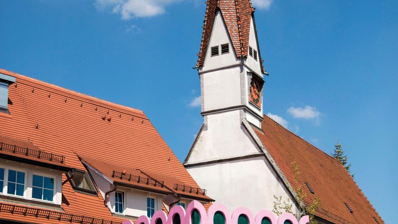 Bunt bemaltes Gebäude mit Drachenmotiv und pinken Details neben einer Kirche mit spitzem Turm in Plochingen., © Stuttgart-Marketing GmbH, Achim Mende