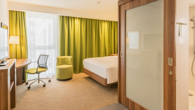 Modernes Hotelzimmer mit Doppelbett, grünem Sessel, Schreibtisch und Blick ins Badezimmer mit Dusche. Helle Vorhänge und grüne Gardinen., © Hampton by Hilton Stuttgart City Centre